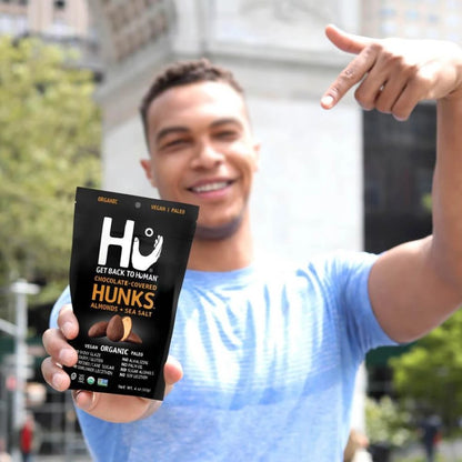 Guy Holding Up And Pointing To Bag Of Hu Hunks Organic Chocolate Covered Almonds